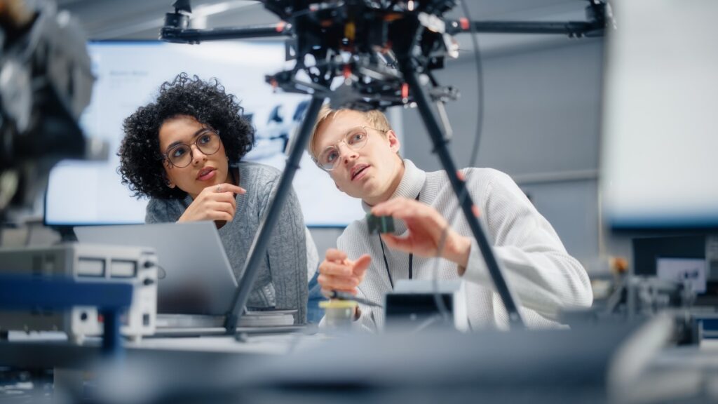 alt= Two people in a lab setting examine a large drone while one holds a component. They are surrounded by various equipment and appear to be discussing the project.